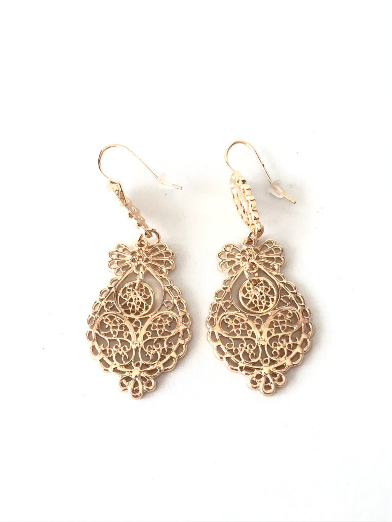 May include: A pair of gold filigree dangle earrings with a delicate, intricate design. The earrings are teardrop shaped and feature a small, round accent at the top.