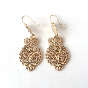 May include: A pair of gold filigree dangle earrings with a delicate, intricate design. The earrings are teardrop shaped and hang from a small hook.