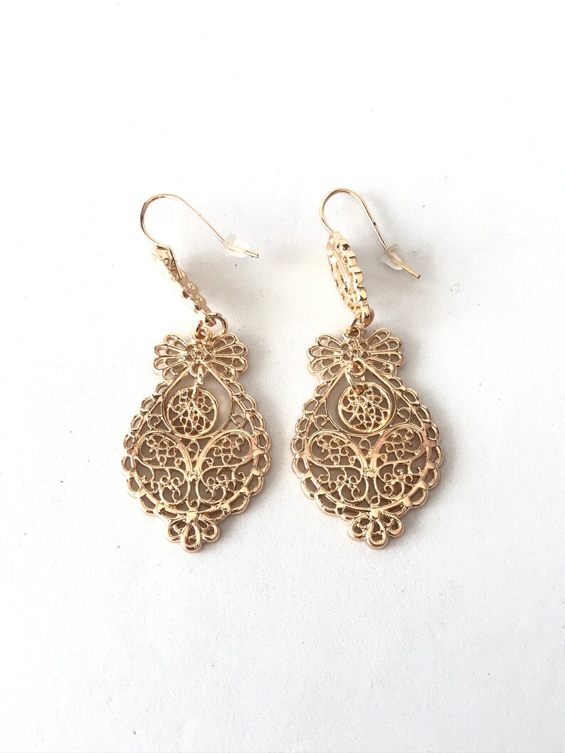 May include: A pair of gold filigree dangle earrings with a delicate, intricate design. The earrings are teardrop shaped and hang from a small hook.