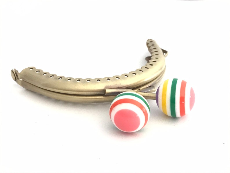 May include: Gold metal purse frame with two colorful striped plastic clasps. The clasps are red, white, green, yellow, and purple.