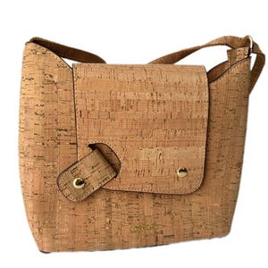 May include: A light brown cork crossbody bag with a flap closure and a gold metal buckle. The bag has a gold metal logo that says "Corkcho".