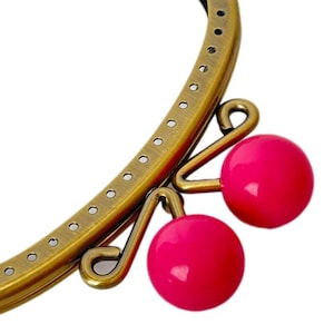 May include: A brass-toned purse frame with a curved design and small holes for sewing. Two bright pink, round beads dangle from the frame. The frame is set against a white background.
