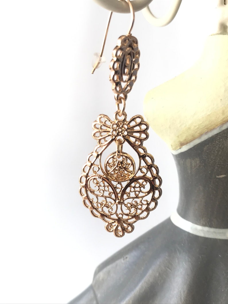 May include: Gold filigree earrings with a bow design. The earrings are dangling and have a delicate, intricate pattern.