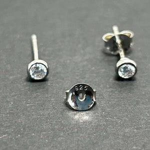 Pode incluir: Um par de brincos de prata com pedras redondas transparentes em cravação bisel. Cada brinco tem um pino. Uma tarracha de prata está incluída, claramente marcada com o selo 925, indicando uma composição de prata de lei.