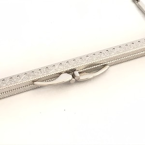 May include: Silver-toned metal purse frame with an ornate design. The frame is closed and the clasp is visible.