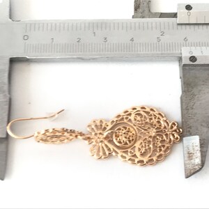 May include: Gold filigree earrings with a delicate design. The earrings are measured with a ruler, showing they are approximately 5 cm long.