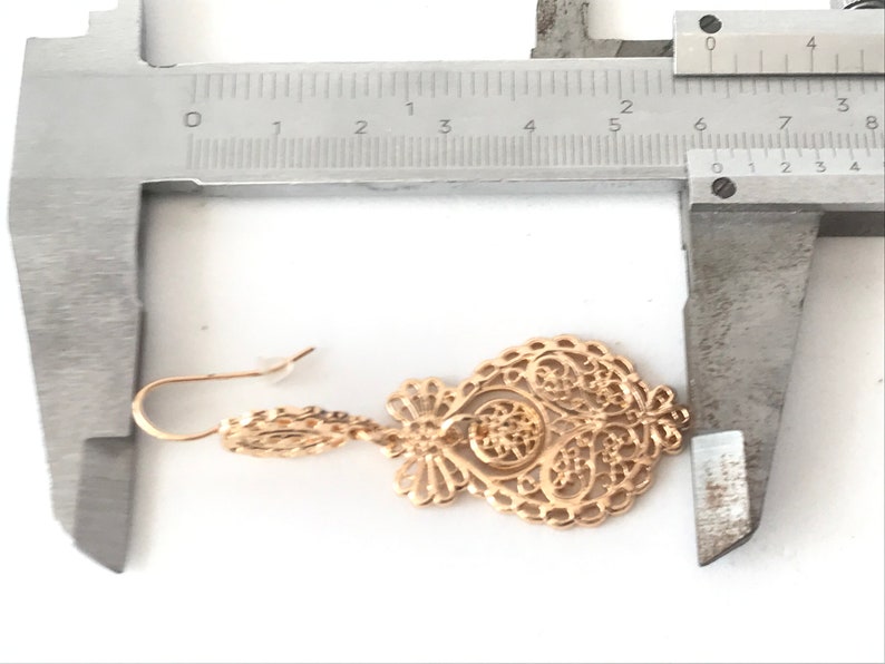 May include: Gold filigree earrings with a delicate design. The earrings are measured with a ruler, showing they are approximately 5 cm long.