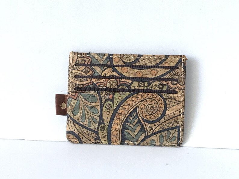 May include: A brown, cork wallet with a paisley pattern in shades of brown, blue, and green. The wallet has multiple card slots and a brown leather pull tab.