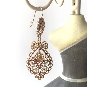 May include: Gold filigree dangle earrings with a bow detail. The earrings are hanging from a white jewellery stand.