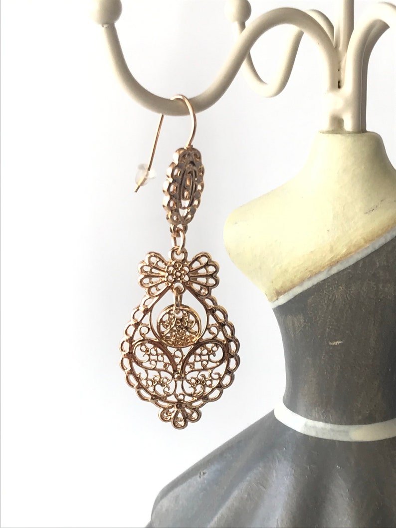 May include: Gold filigree dangle earrings with a bow detail. The earrings are hanging from a white jewellery stand.