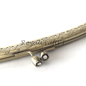 Può includere: Cornice per borsa in ottone antico con dettagli floreali ornati. La cornice presenta un design curvo con abbellimenti decorativi e due piccoli accenti a forma di gioiello. Il testo "RoyalcraftPT" è visibile.