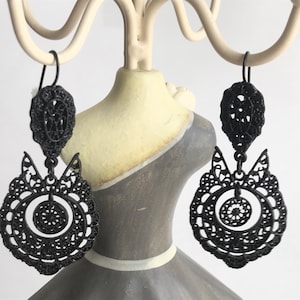 Può includere: Un paio di orecchini pendenti neri con un design delicato e intricato. Gli orecchini presentano un motivo circolare con un cerchio più piccolo al centro.