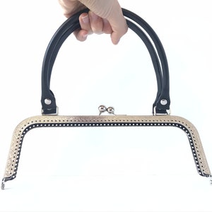 Pode incluir: Uma armação de bolsa com uma alça preta e um fecho prateado. A armação tem uma borda decorativa com pequenos orifícios. A alça é fixada à armação com rebites prateados. A armação da bolsa é segurada por uma mão em um fundo branco.