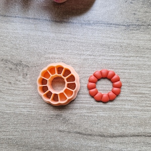 Peut inclure: Deux emporte-pièces en forme de fleur de couleur orange. L'un a un design détaillé avec des découpes intérieures, tandis que l'autre est un simple anneau de petites formes arrondies. Les emporte-pièces sont sur une surface grise.