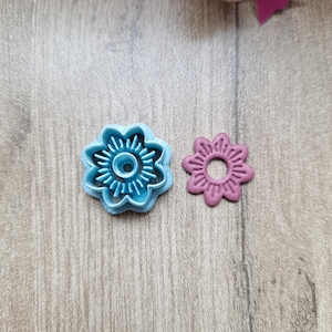 Peut inclure: Deux emporte-pièces en forme de fleur. L'un est bleu clair, l'autre rose. Le découpoir bleu a un centre détaillé et des pétales extérieurs. Le découpoir rose est une forme pleine avec un trou au centre. Sur une surface en bois.