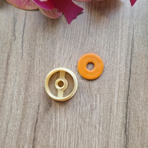Peut inclure: Deux emporte-pièces : un beige en forme de roue avec un trou central et des rayons, et un orange, circulaire avec un trou au milieu. Les emporte-pièces sont sur une surface en bois, avec des pétales d'orchidée roses et violets en arrière-plan.