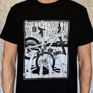Könnte beinhalten: Schwarzes T-Shirt mit einer weißen Grafik eines Mountainbikes, Werkzeugen und den Worten "DIY or DIE" und "RIDE & DIE". Das Fahrrad steht im Mittelpunkt, mit Werkzeugen und einer Reparaturwerkstatt im Hintergrund.