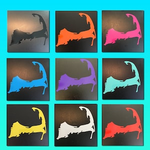 May include: Nine vinyl stickers featuring the outline of Cape Cod in different colors, including black, orange, pink, blue, purple, turquoise, yellow, white, and red. The stickers are on a black background.