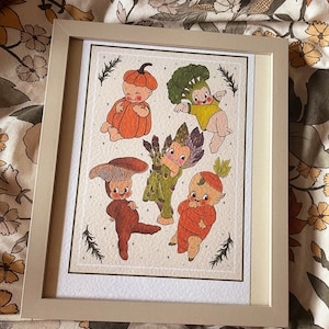 May include: A framed illustration of five cartoon babies dressed as vegetables. The babies are dressed as a pumpkin, broccoli, mushroom, asparagus, and carrot. The illustration is on a white background with green leaves and white stars.