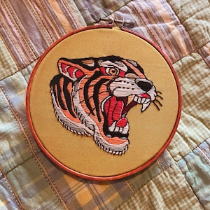 May include: Embroidered tiger head in a wooden hoop with a beige background. The tiger is facing right and has a red open mouth with white teeth. The tiger has black stripes and orange and white fur.