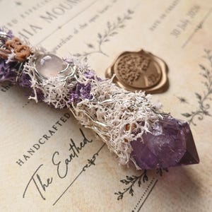 May include: A decorative wand featuring a deep purple amethyst crystal point at one end, wrapped in white reindeer moss and silver wire. The handle is adorned with a smooth, pale pink gemstone and small brown floral accents, creating a textured look.