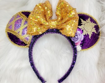 May include: Purple and gold mouse ears with a gold bow, a sun design, and purple flowers.