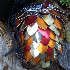 Peut inclure: Un sac à dés en métal doré, rouge et orange avec une chaîne et un cordon de serrage en cuir noir. Le sac est fait d'écailles et repose sur un lit de feuilles brunes.