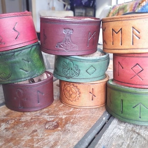 May include: A stack of eight leather bracelets with various designs and symbols, including a dragon, a hammer, runes, and a gear. The bracelets are in shades of brown, red, and green.