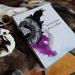 K&ouml;nnte beinhalten: Das illustrierte Buch der Runen, ein wei&szlig;es Buch mit einem schwarzen und violetten Aquarelldesign, das einen Kreis von Runen zeigt. Das Buch liegt auf einer braunen Pelzoberfl&auml;che.