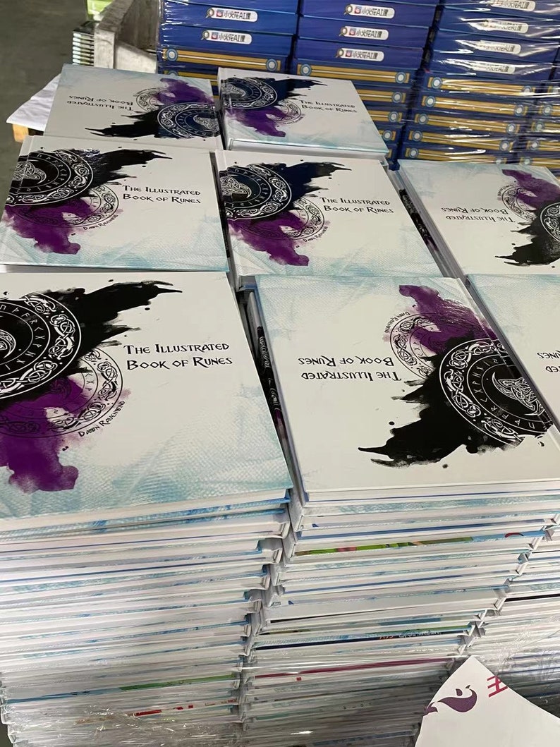 K&ouml;nnte beinhalten: Ein Stapel wei&szlig;er B&uuml;cher mit einem blauen und wei&szlig;en Aquarelldesign auf dem Cover. Der Titel des Buches lautet "The Illustrated Book of Runes".