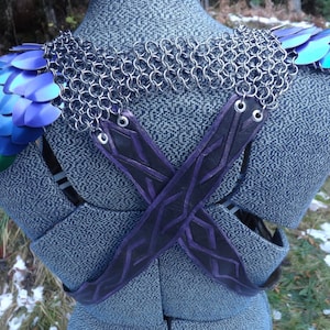 May include: A fantasy shoulder piece featuring rainbow-coloured scales and silver chainmail. It has black and purple leather straps that cross at the back. The shoulder armour is displayed on a grey tweed mannequin.