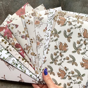 May include: A collection of stationery envelopes featuring various nature-inspired patterns, including illustrations of squirrels, pinecones, pine branches, deer, flowers, acorns, and mushrooms on off-white paper with diverse color palettes.