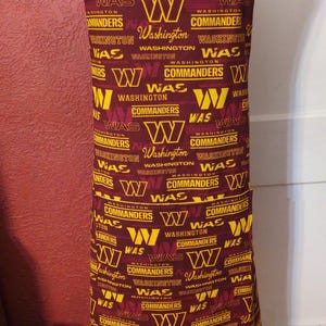 Op de afbeelding: Een bordeauxrode schort met het logo en de naam van de Washington Commanders in goud. De schort is bedrukt met het logo en de naam van het team en heeft een zak. De schort wordt op een mannequin getoond.
