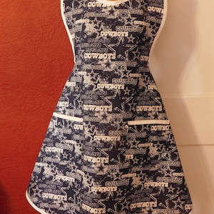 May include: A blue and white apron with a white trim and a pattern of stars and the word "Cowboys" repeated throughout.