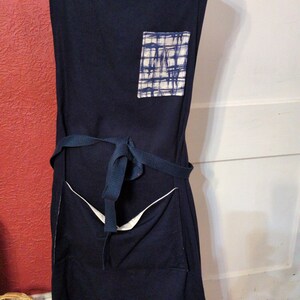 May include: A navy blue apron with white accents and a pocket featuring a blue and white plaid pattern. The apron has a tie-waist design and adjustable straps with a matching plaid pattern.