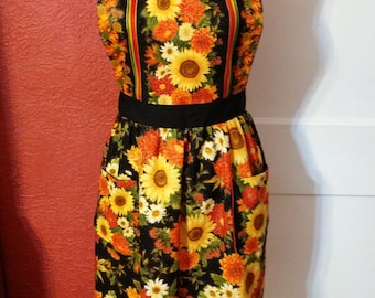 May include: A black apron with a sunflower and floral print. The apron has a black waistband and two pockets.