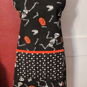 May include: Black Halloween apron with a white skeleton and orange pumpkin pattern. The apron has a white trim, an orange pocket border, and black straps with a skull pattern. A fun and festive kitchen accessory.