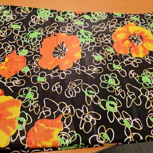 May include: Black fabric with a repeating pattern of orange and yellow flowers with green centers. The background is black with white and green swirls.