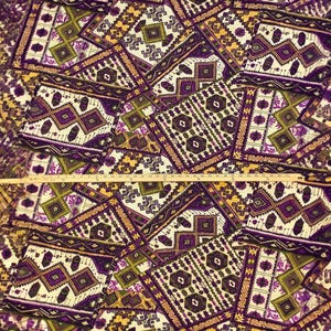 May include: Geometric patterned fabric in shades of purple, white, olive green, and gold. The design features repeating diamond and rectangular shapes with intricate details. A wooden ruler is placed across the fabric, measuring approximately 24 inches.