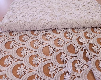 May include: White lace fabric with a floral and scalloped design. The fabric is folded over, revealing the intricate pattern. The lace is a delicate, open weave, perfect for apparel or home decor projects.