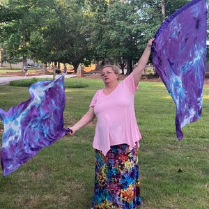 May include: A woman holds two large, flowing flags with a tie-dye pattern of purple, blue, and white. The flags are held up in the air against a backdrop of green grass and trees. The woman is wearing a pink top and a colorful skirt.