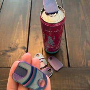 May include: A collection of iridescent can openers in various shapes and sizes. One opener is attached to a pink can of "Pink Slush" energy drink. The openers have a key ring hole and a textured surface.