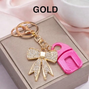 May include: Gold-colored keychain with a bow-shaped charm encrusted with rhinestones and a pink accessory. The word "GOLD" is printed in black at the top. The keychain is attached to a silver-colored ring.
