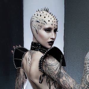 May include: A woman with a shaved head and a black dress with a tulle skirt. She has a studded collar and shoulder pads. Her head is covered in spikes and she has tattoos on her arms.