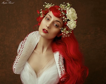 May include: A woman with long red hair wears a white and red corset with a white ruffled collar and a white tulle skirt. She is wearing a floral crown with white roses.