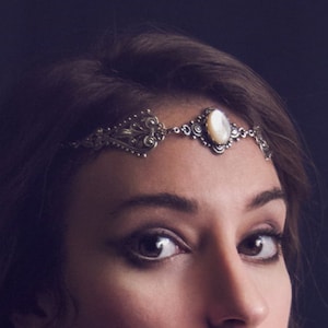 May include: A woman with brown hair and red lipstick wears a silver headband with a central oval pearl. The headband has an ornate, vintage design. The background is a dark, solid colour.