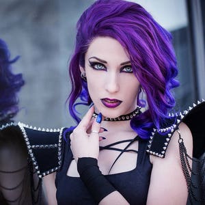 May include: A woman with long, bright purple hair wears a black top with a criss-cross design and a studded black leather shoulder piece. She is wearing a black choker with silver studs and has dark eye makeup.