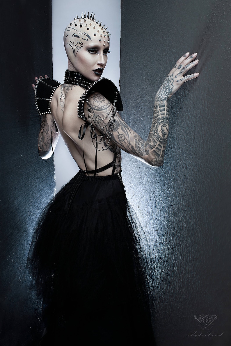 May include: A woman with a shaved head and a black dress with a tulle skirt. She has a studded collar and shoulder pads. Her head is covered in spikes and she has tattoos on her arms.