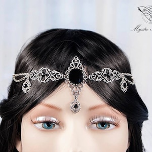 Pode incluir: Uma tiara prateada com pedras pretas. O design ornamentado apresenta uma grande pedra central e pequenos detalhes, com pingentes pendurados em forma de gota. O logotipo Mystic Thread é visível.