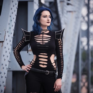 May include: A woman with blue hair is wearing a black, cut-out bodysuit with studded shoulder pads. She is standing in front of a metal structure.